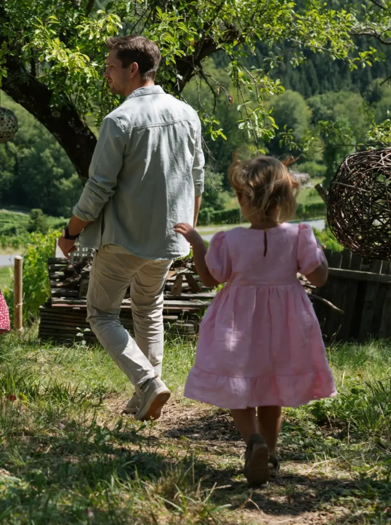 Ein Erwachsener und ein Kind gehen gemeinsam durch eine grüne Landschaft, umgeben von Bäumen und Natur.