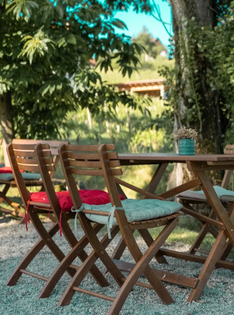 Holztisch mit bunten Stühlen in einem sonnigen Garten, umgeben von Bäumen und sanften Hügeln.