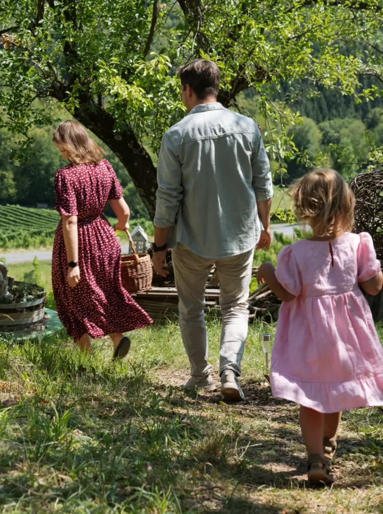 Eine Familie spaziert durch einen Garten, umgeben von Bäumen und Weinbergen, mit einem Korb in der Hand.